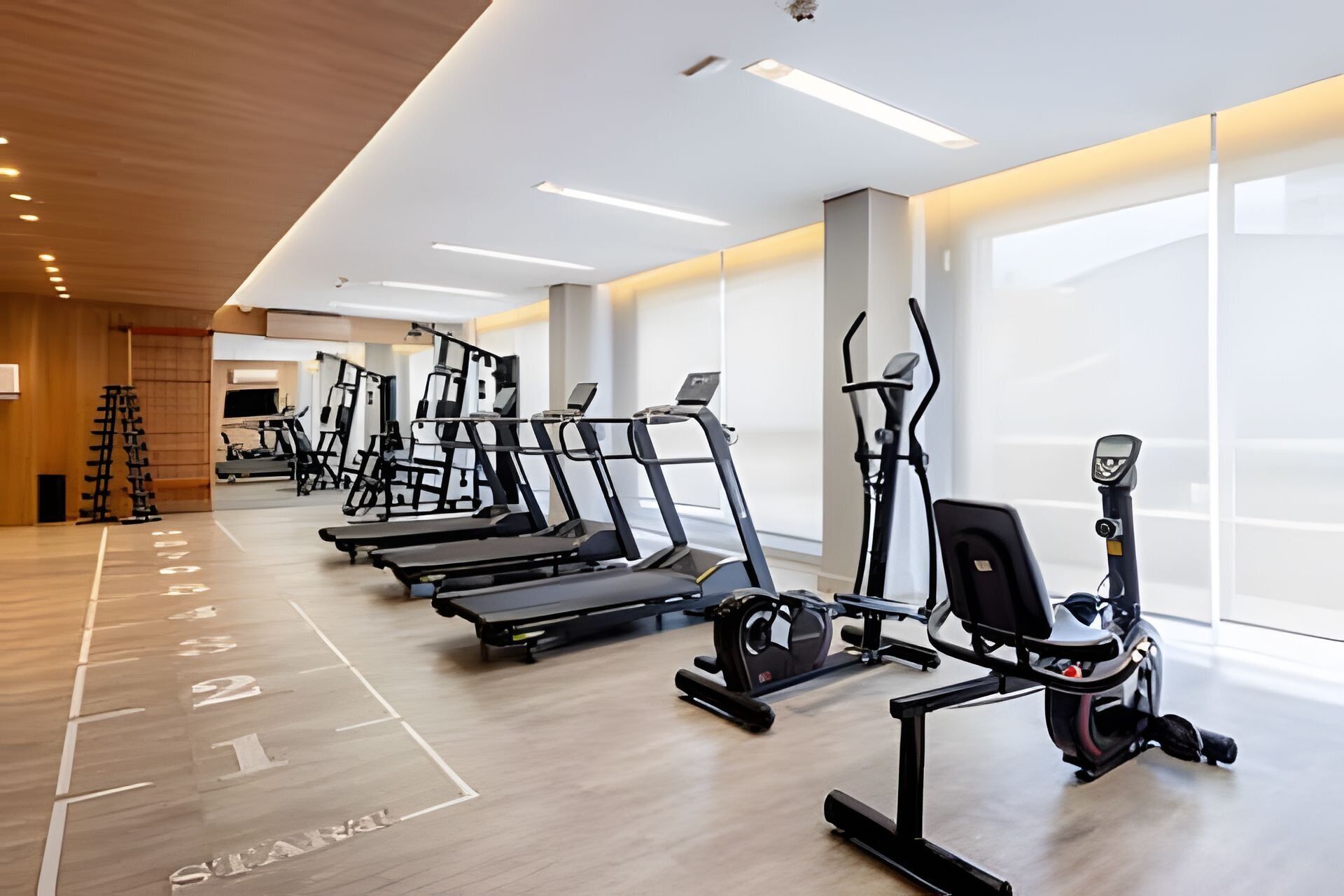 Uma academia moderna com vários equipamentos de cardio, piso de madeira e janelas amplas.