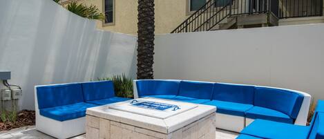 An outdoor seating area with a stone fire pit, blue cushioned sofas, and a white wall.