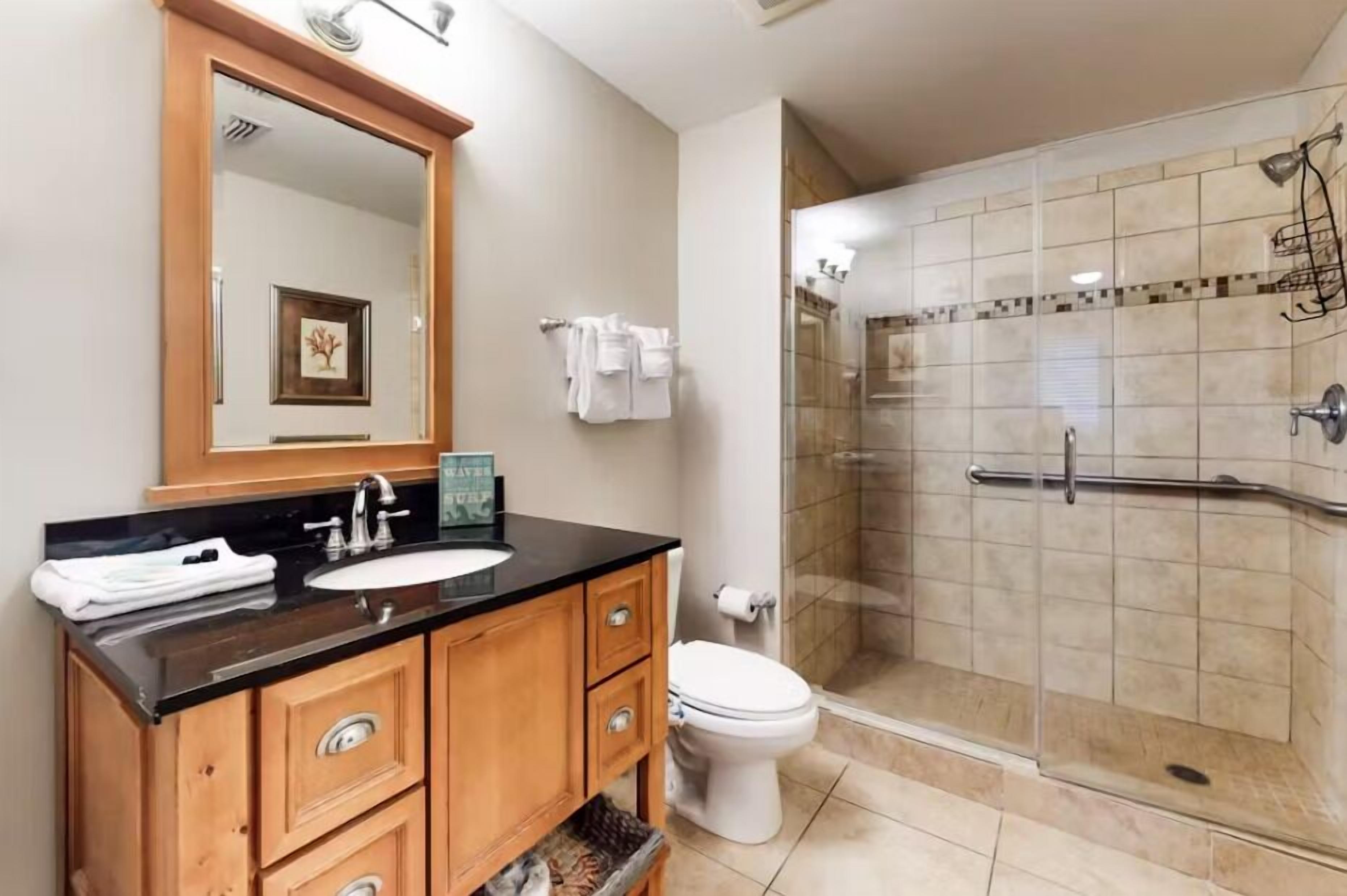 Une salle de bain moderne avec un plan de toilette noir, des meubles en bois, un lavabo blanc et une douche séparée par une paroi de verre.