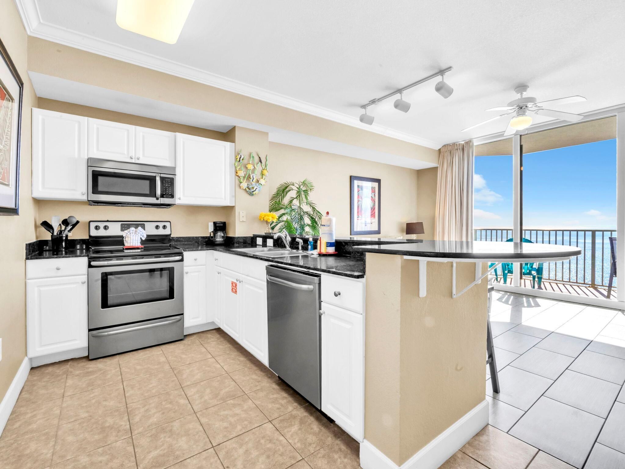 Cocina moderna con armarios blancos, microondas, estufa y lavavajillas. Hay una pequeña isla con un lavaplatos y vistas al balcón, que cuenta con barandilla y sillas.