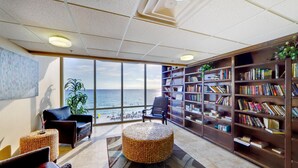 Een bibliotheek met een groot raam met uitzicht op zee, een zwarte leren bank en een boekenplank vol boeken.