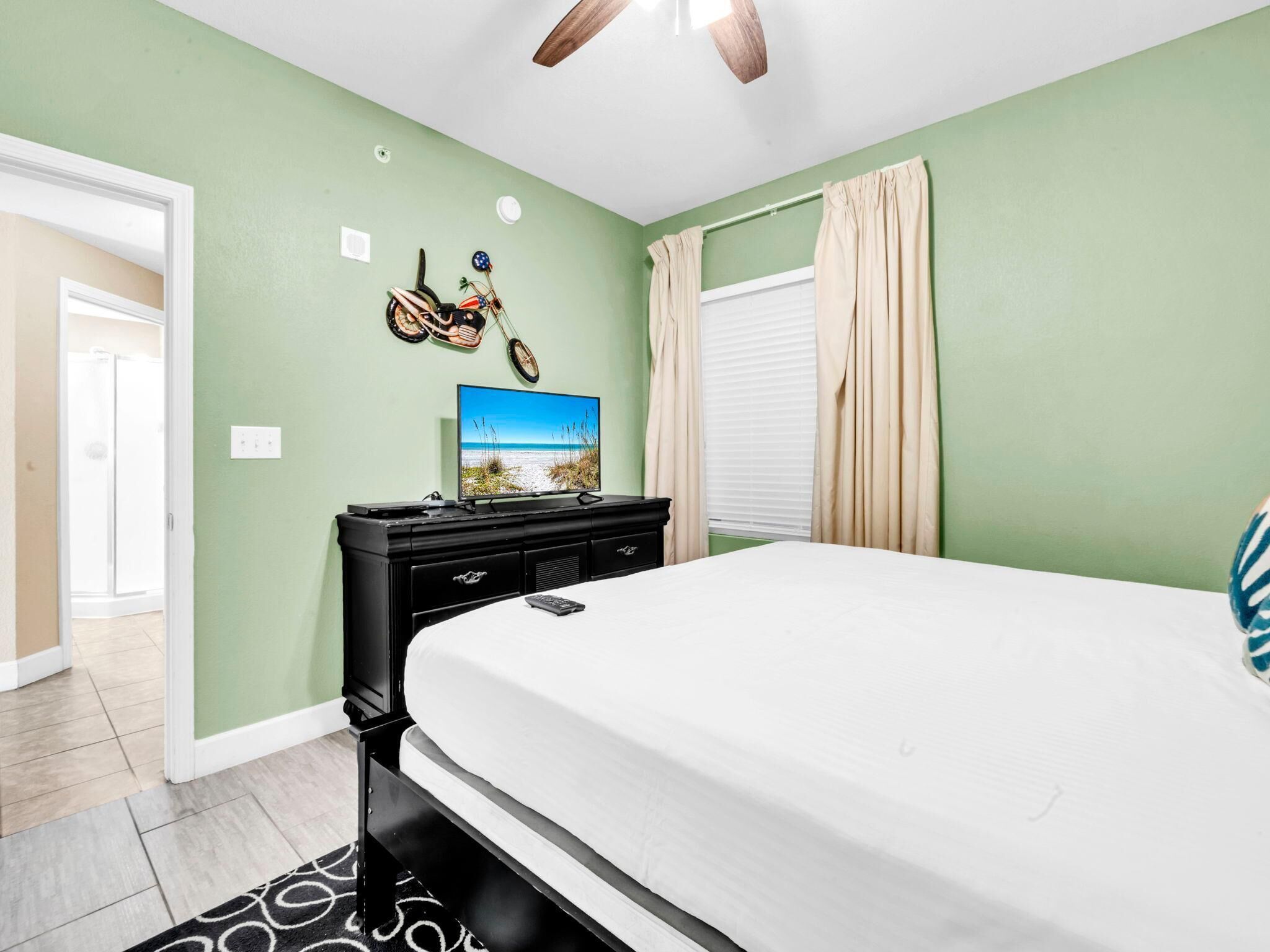 A bedroom with a bed, a dresser, a television on the wall, and a window with curtains.
