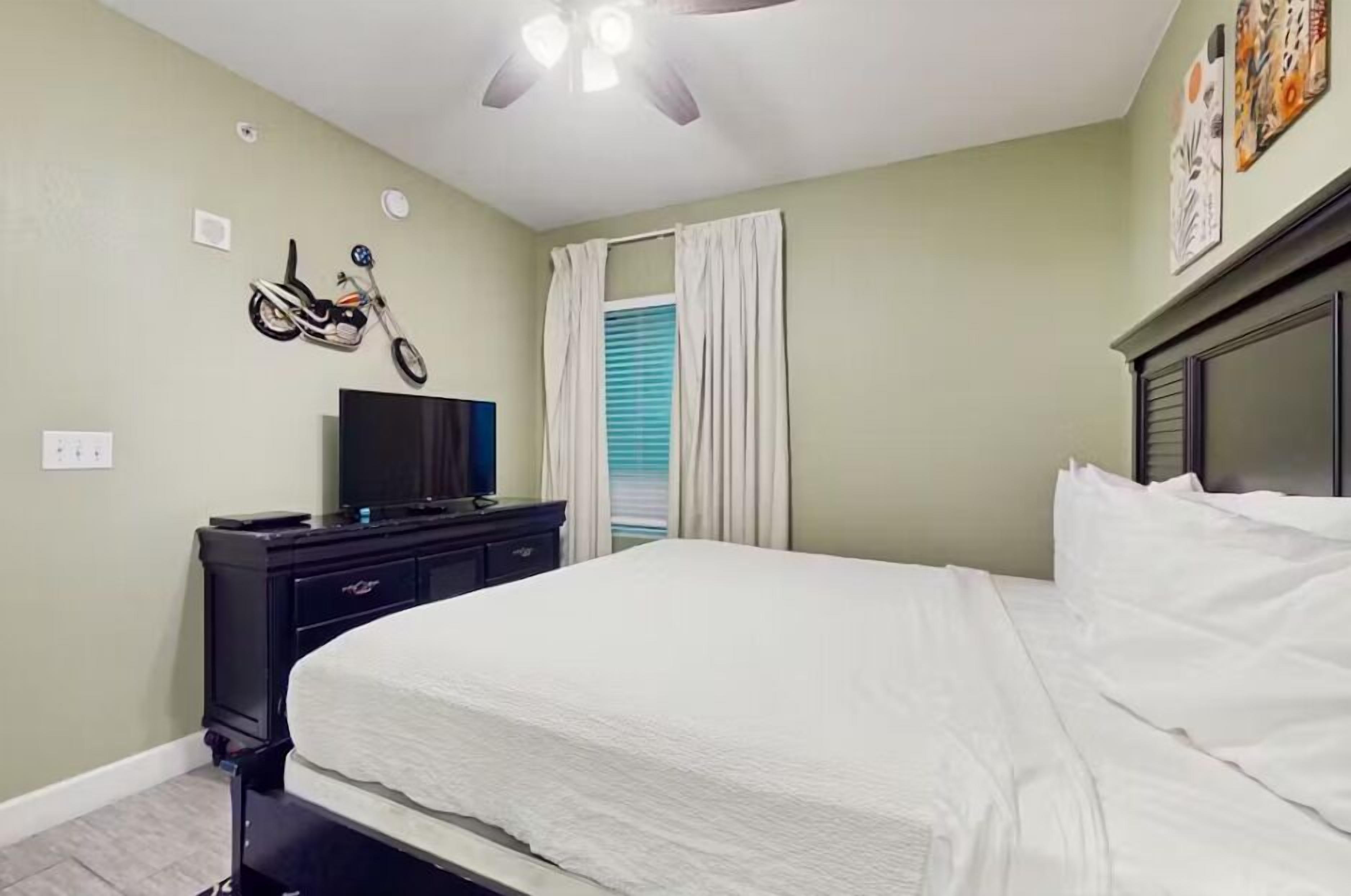 A bedroom with a bed, a dresser, a television, a ceiling fan, and a window with curtains.