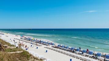 A beach with rows of striped beach umbrellas and lounge chairs, a pier in the background, and a clear blue sky.