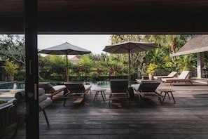 Um deck de madeira com cadeiras de lounge e guarda-sóis, com vista para uma piscina e vegetação exuberante.