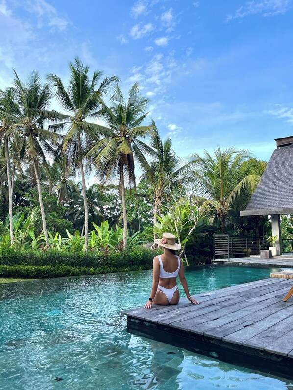 A person sitting on a wooden dock by a clear pool surrounded by palm trees and lush greenery. - Luxury 6-Bedroom Villa near Ubud - Private Pool, Nature & Privacy (Ubud)