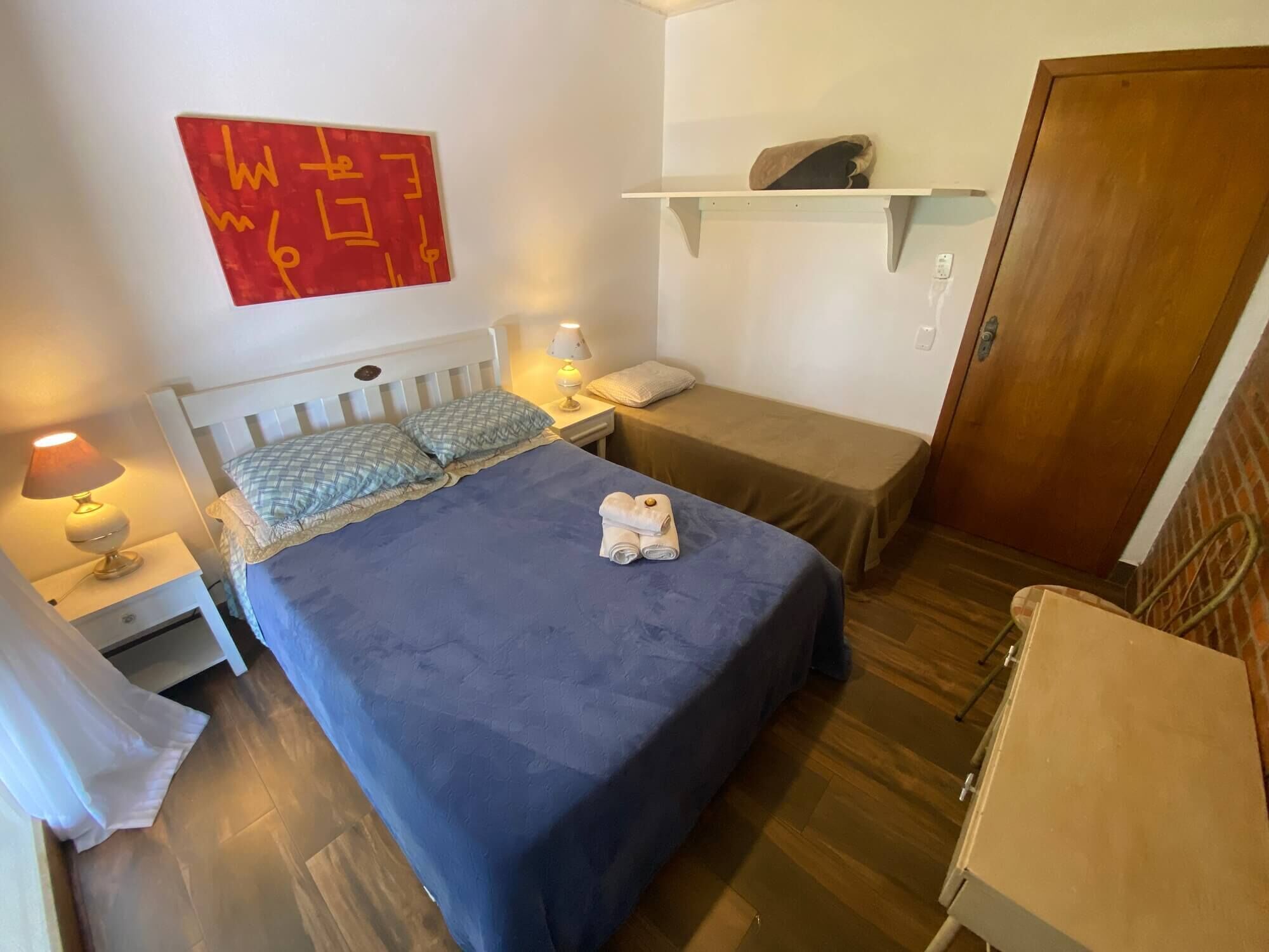 A room with a bed, a bedside table with a lamp, a small shelf, and a door.