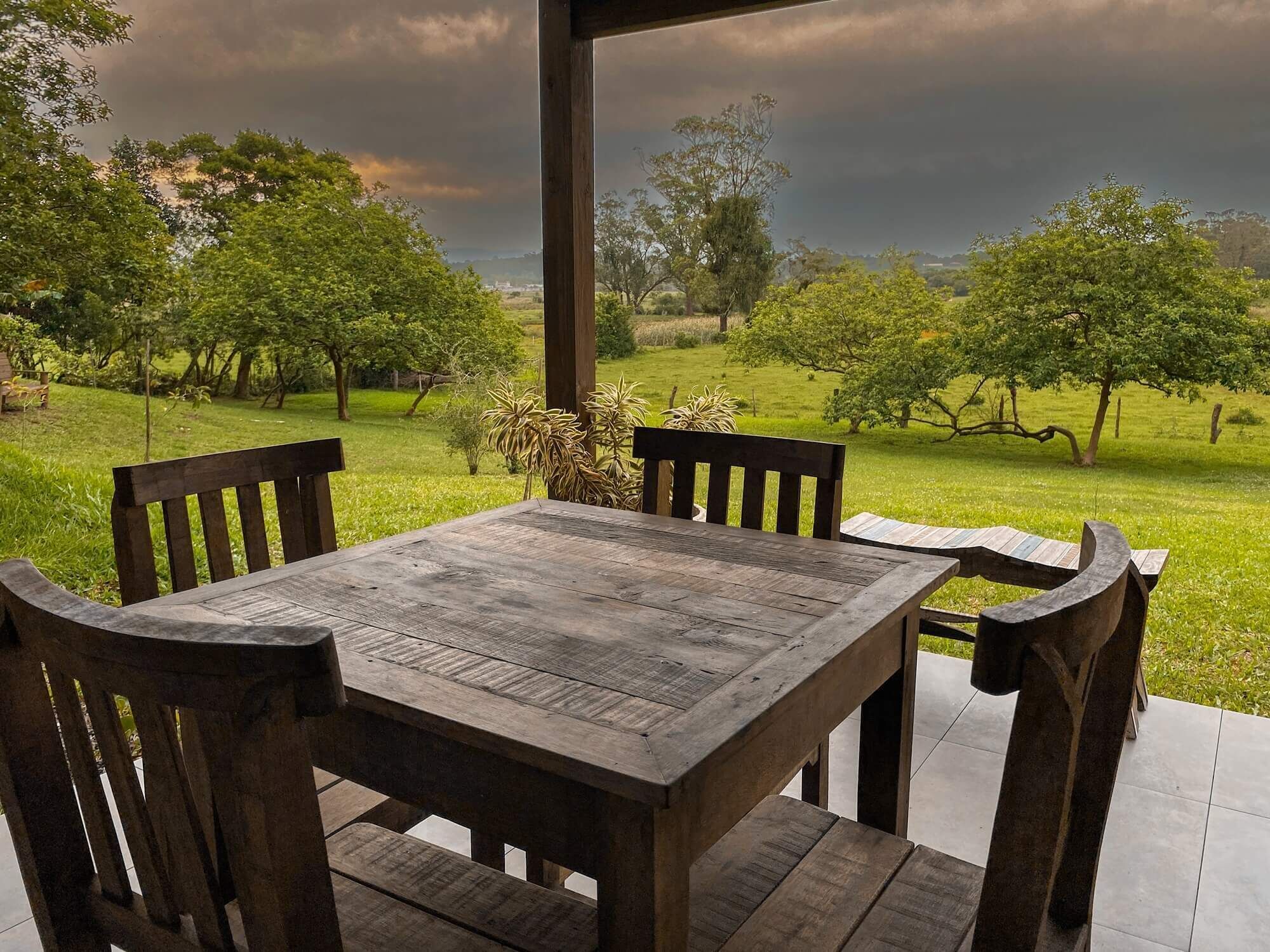A wooden table and chairs set on a patio with a view of a grassy landscape and trees.
