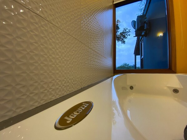 A bathtub with a Jacuzzi logo, a textured wall, and a window with a view of a building and trees. - Morada das Bromélias (Torres)