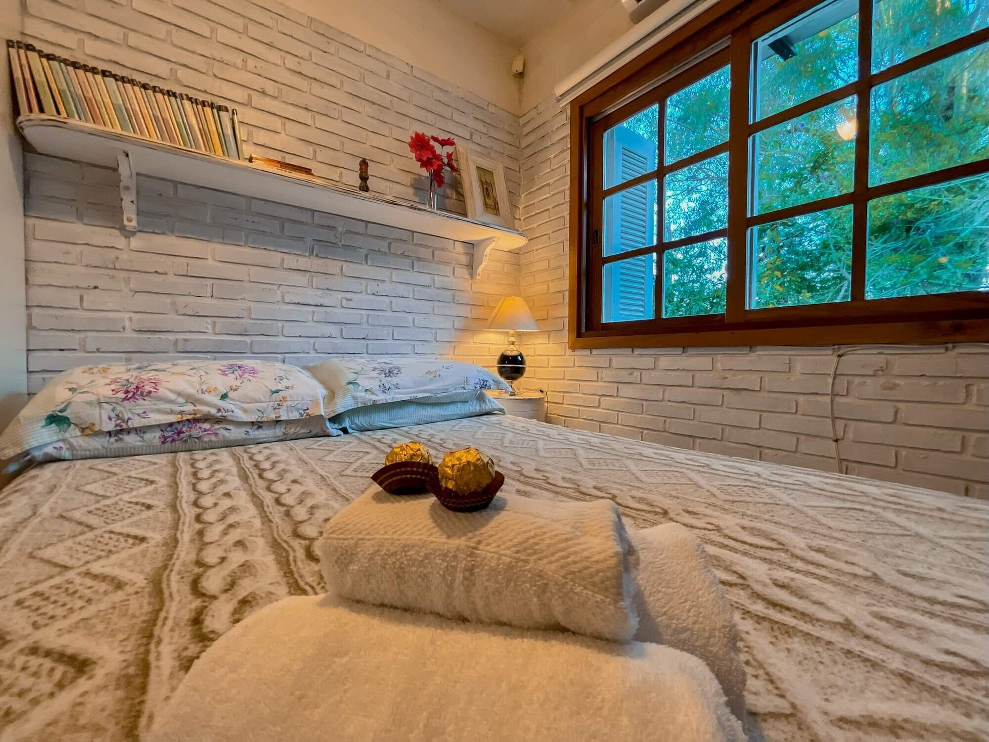 A cozy bedroom with a brick wall, a large window with a view of trees, a bed with floral bedding, and a shelf with books and a vase of flowers.