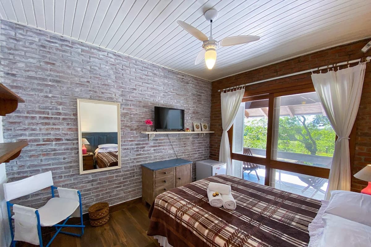 Une chambre avec un mur en briques, un lit recouvert d’une couverture à motifs, un téléviseur posé sur une étagère, un ventilateur de plafond et une fenêtre avec des rideaux.