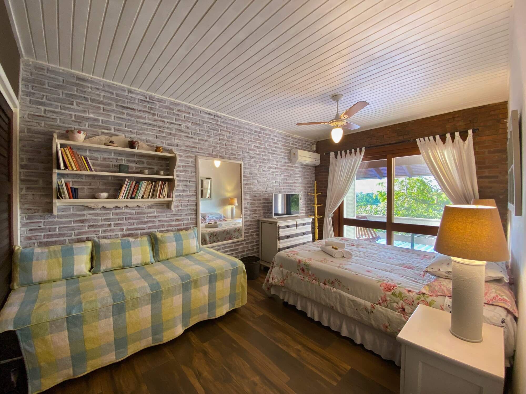 A cozy bedroom with a brick wall, a ceiling fan, a bed with floral bedding, a sofa with checkered upholstery, and a bedside lamp.