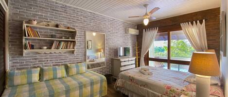 Ein gemütliches Schlafzimmer mit Backsteinwand, Deckenventilator, einem Bett mit floralem Bettzeug und einem Sofa mit kariertem Bezug sowie einer Nachttischlampe.