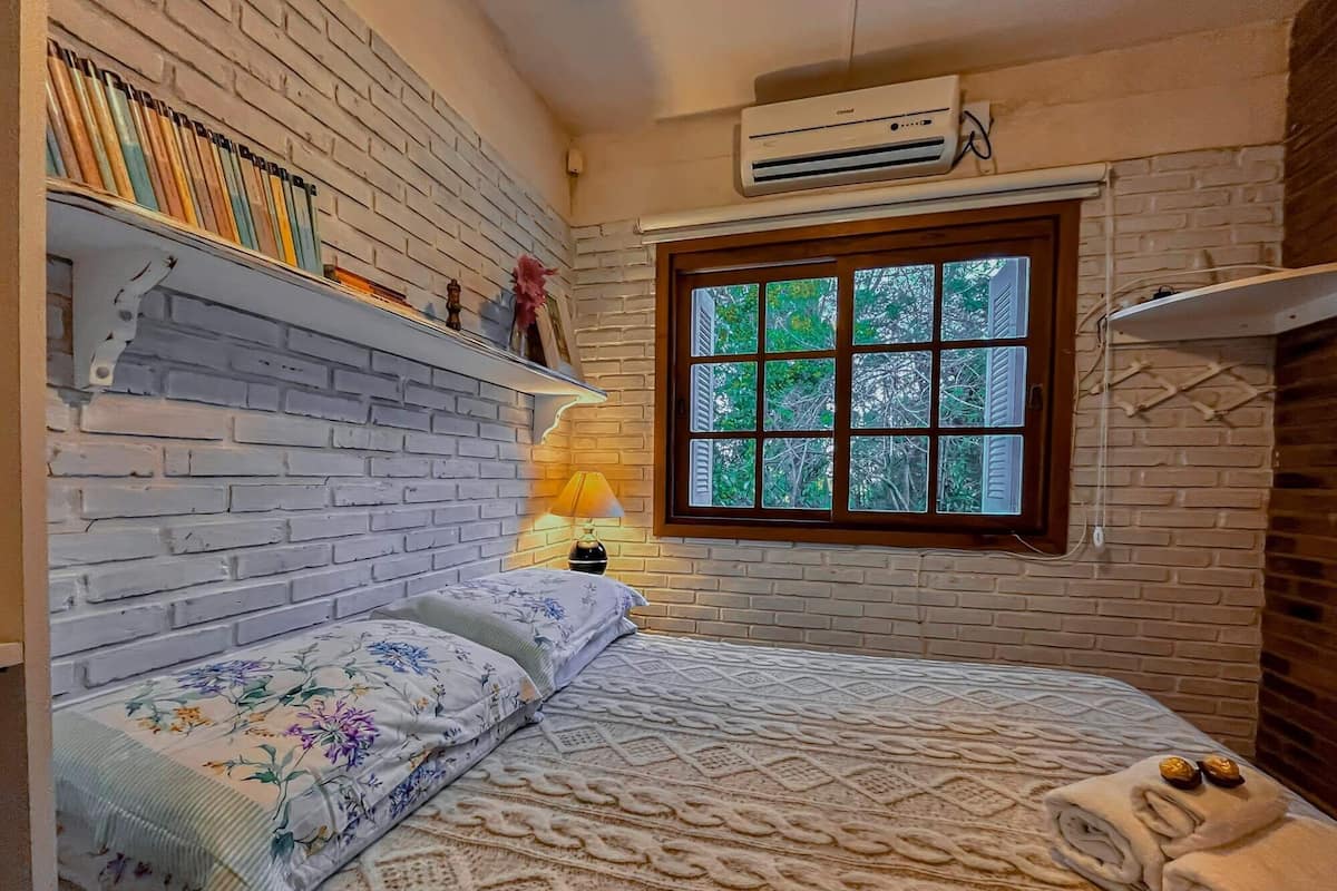 Une chambre avec un mur en briques, une fenêtre donnant sur des arbres, un lit avec une literie à motifs floraux et une étagère remplie de livres.