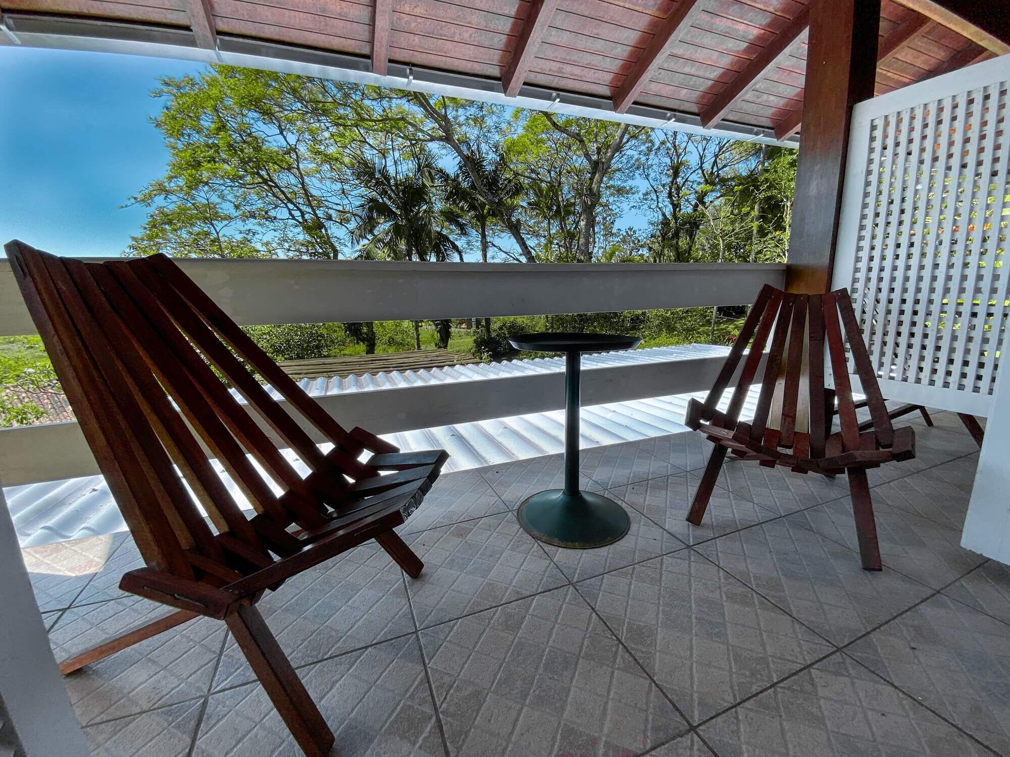 A covered outdoor seating area with two wooden chairs and a small table, overlooking a garden with trees and a clear sky.