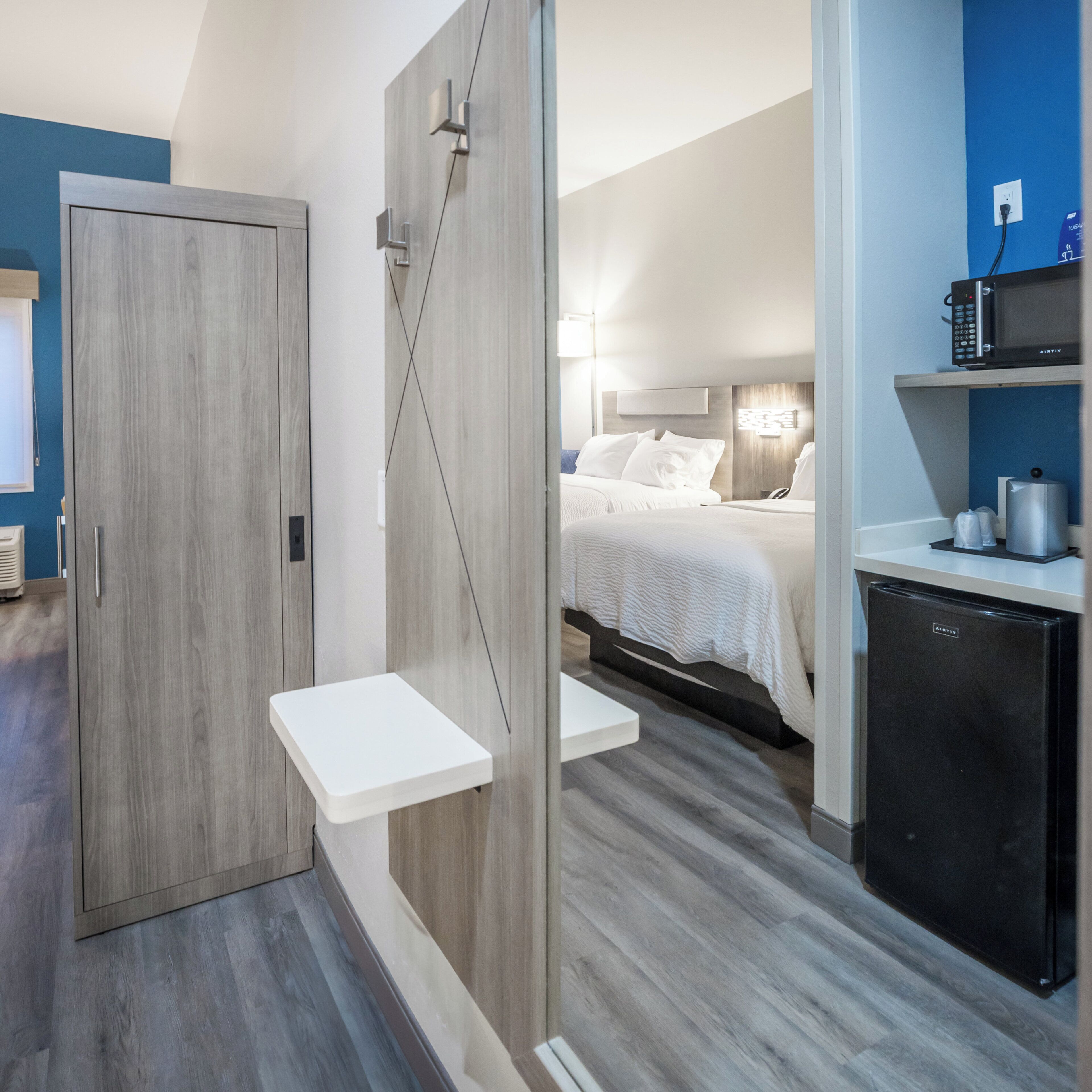 Moderni hotellihuone, jossa on puinen vaatekaappi, yöpöytä, valkoisilla vuodevaatteilla varustettu sänky, mikroaaltouuni ja minijääkaappi.