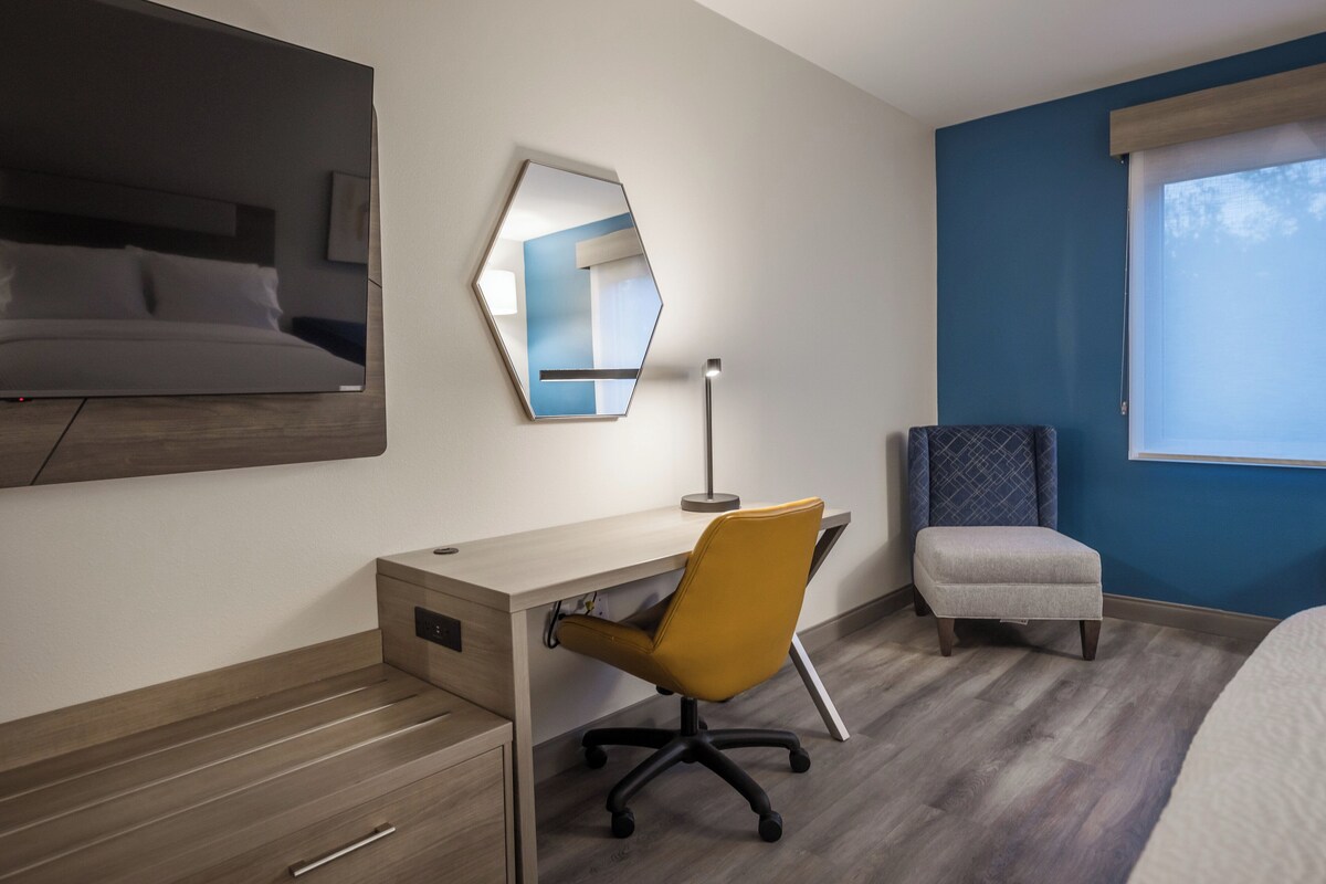 Ein Hotelzimmer mit einem Bett, einem Schreibtisch mit Stuhl, einem großen Spiegel und einem Fenster mit Vorhängen.
