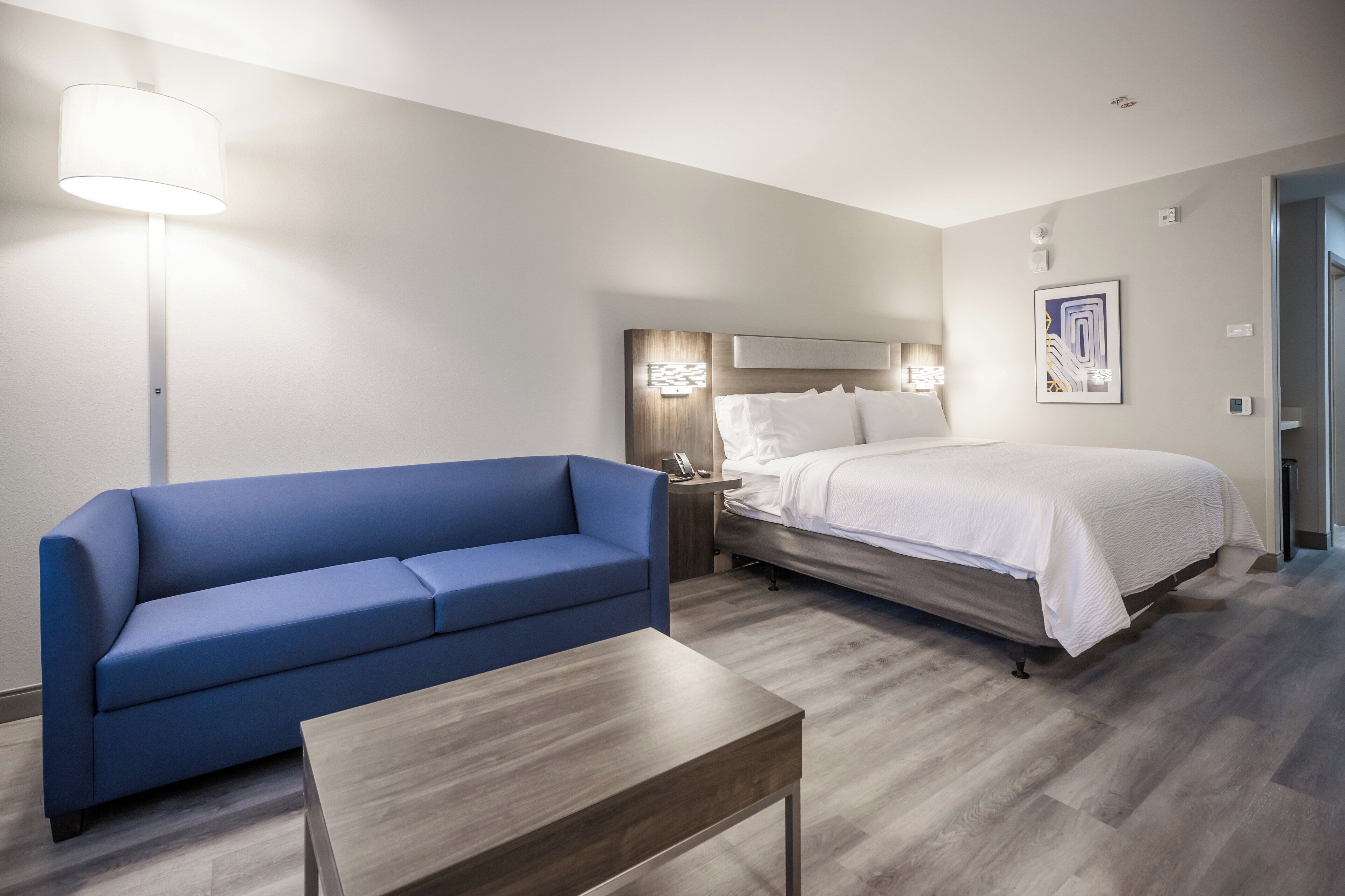 Una habitación de hotel con una cama, un sofá azul, una mesita pequeña y una mesita de noche con una lámpara.