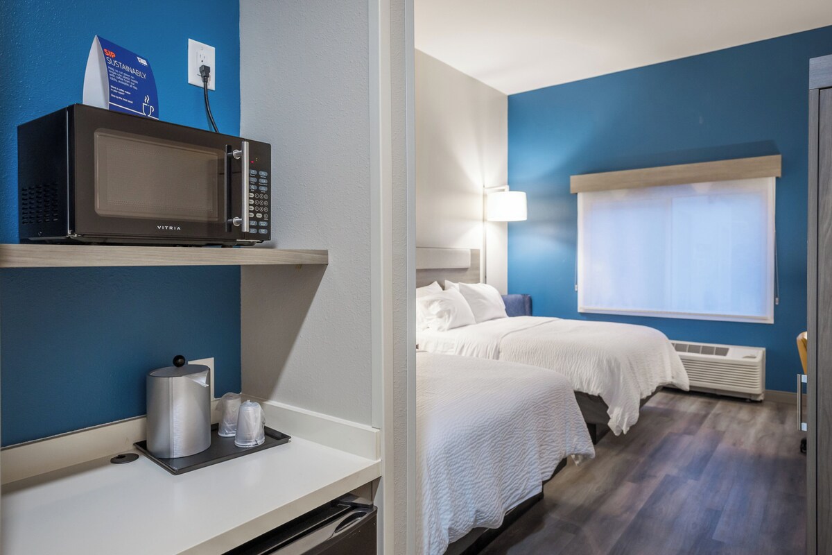 A hotel room with a microwave on a shelf, a bed with white bedding, a bedside lamp, and a window with blinds.