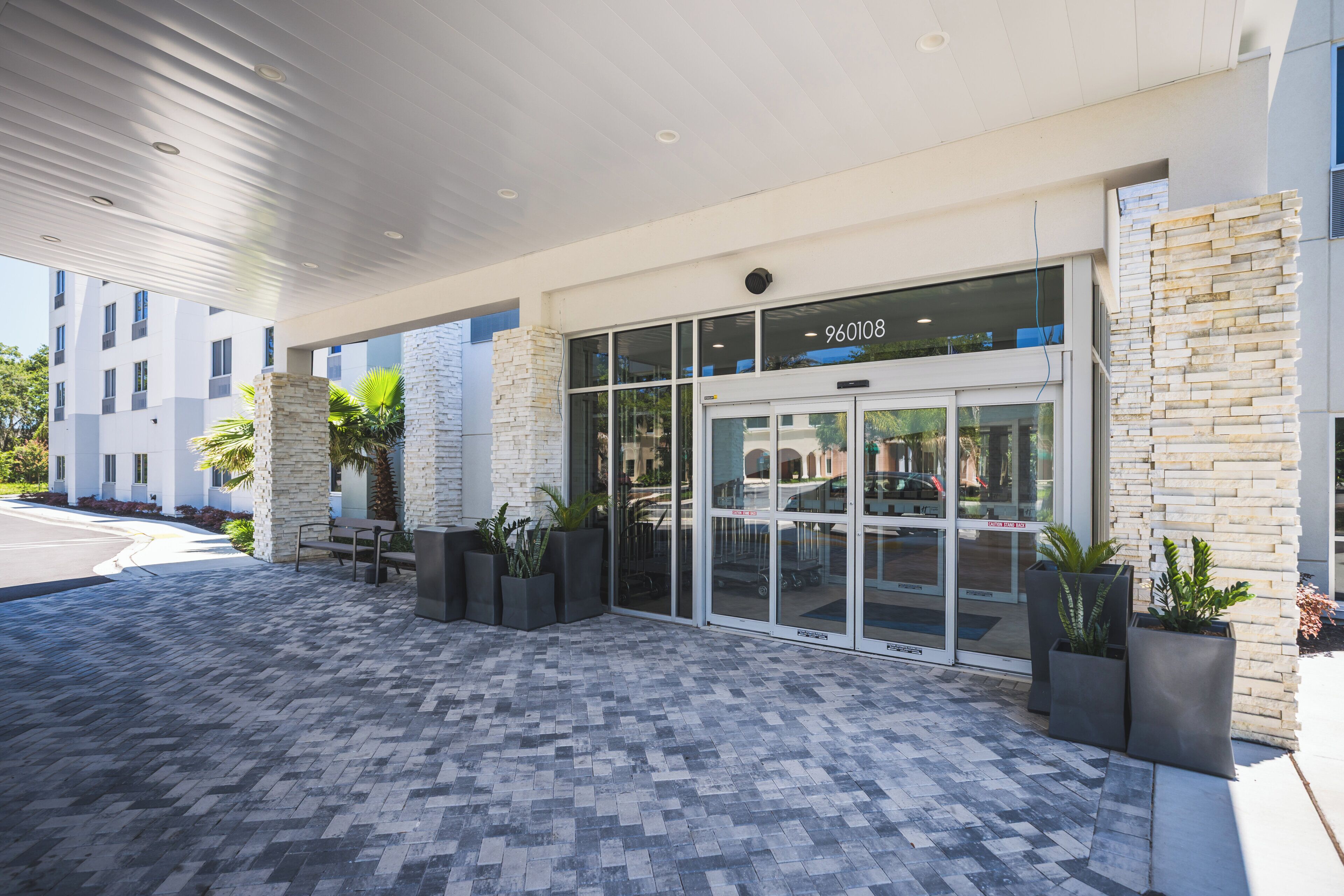 A hotel entrance with a modern design, featuring a glass door with a number sign, a paved walkway, and potted plants.