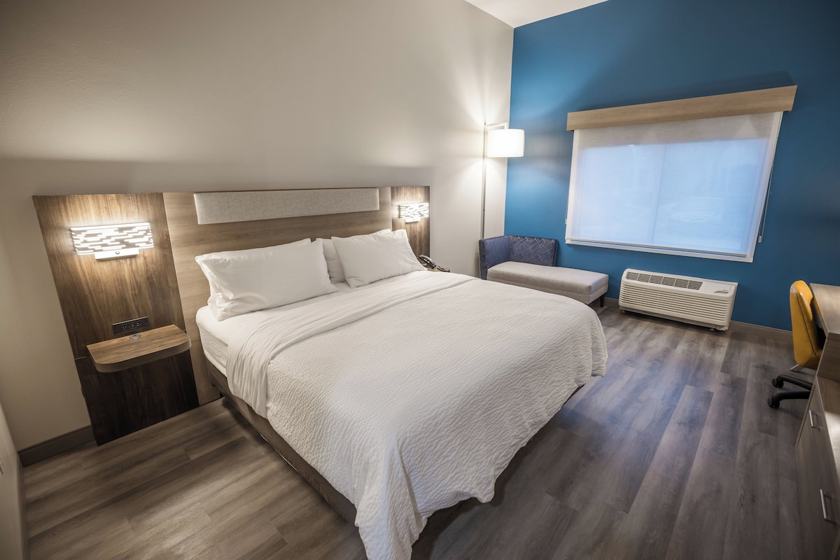 A hotel room with a large bed, bedside tables, a window with blinds, a wall-mounted heater, and a small seating area.