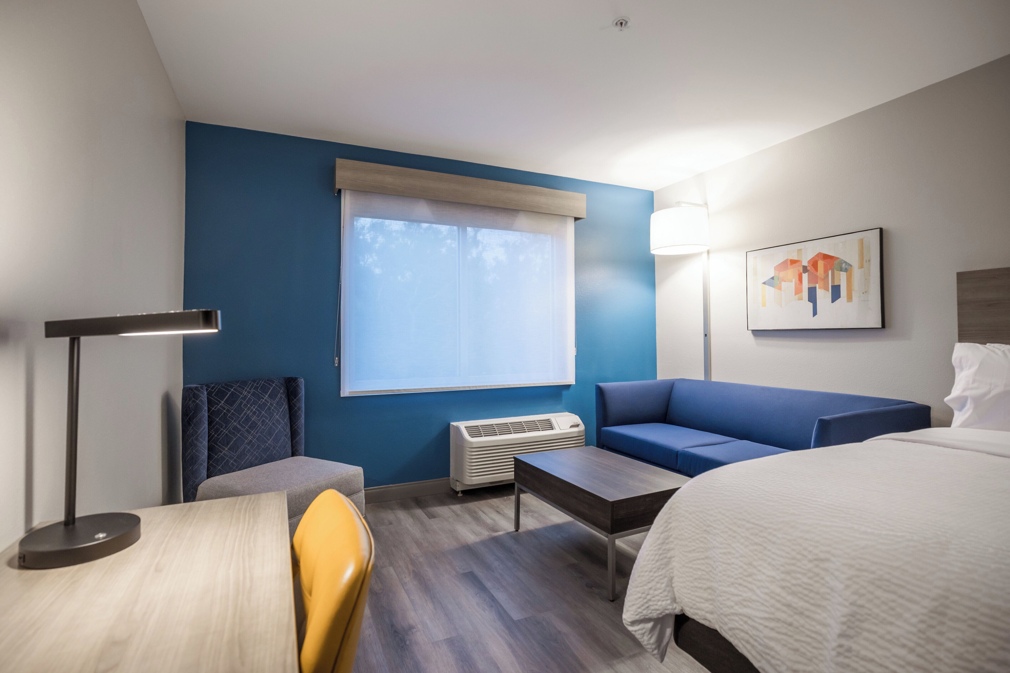 A hotel room with a bed, a sofa, a desk, a chair, a lamp, a window with blinds, and a wall-mounted air conditioner.