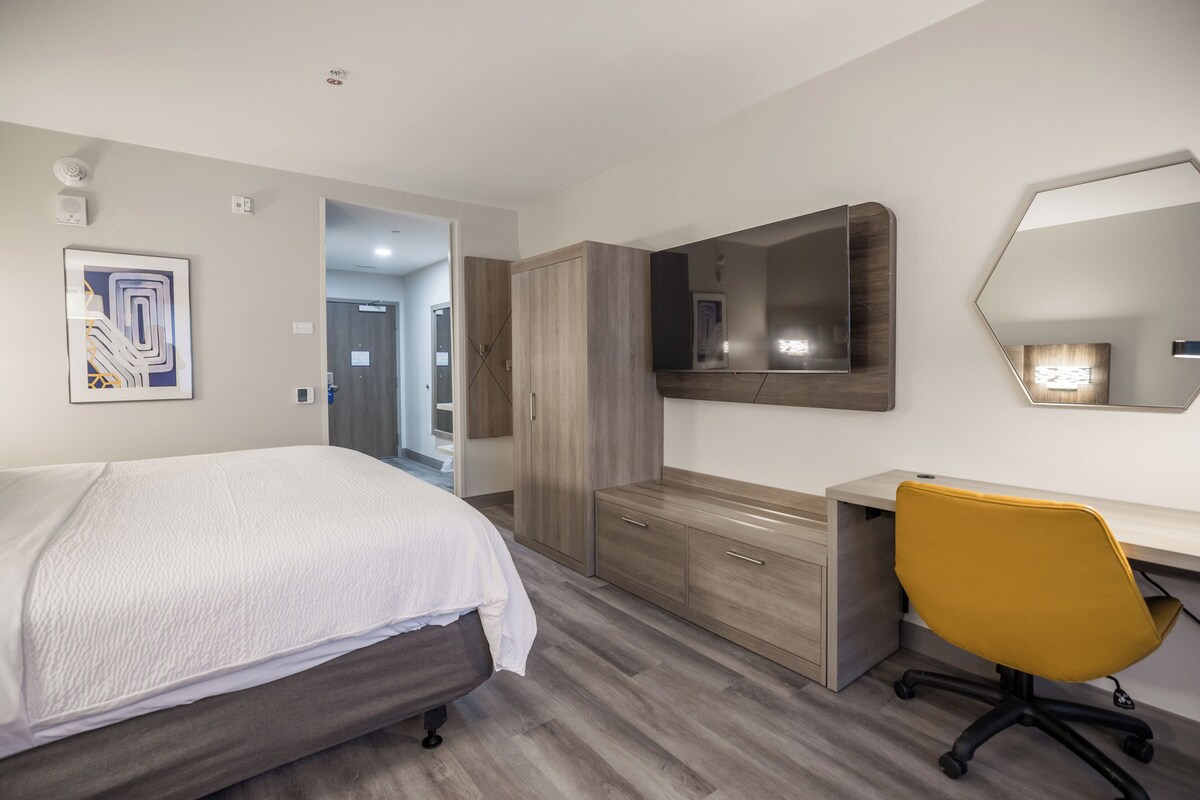 A modern hotel room with a bed, a desk, a chair, and a large mirror.