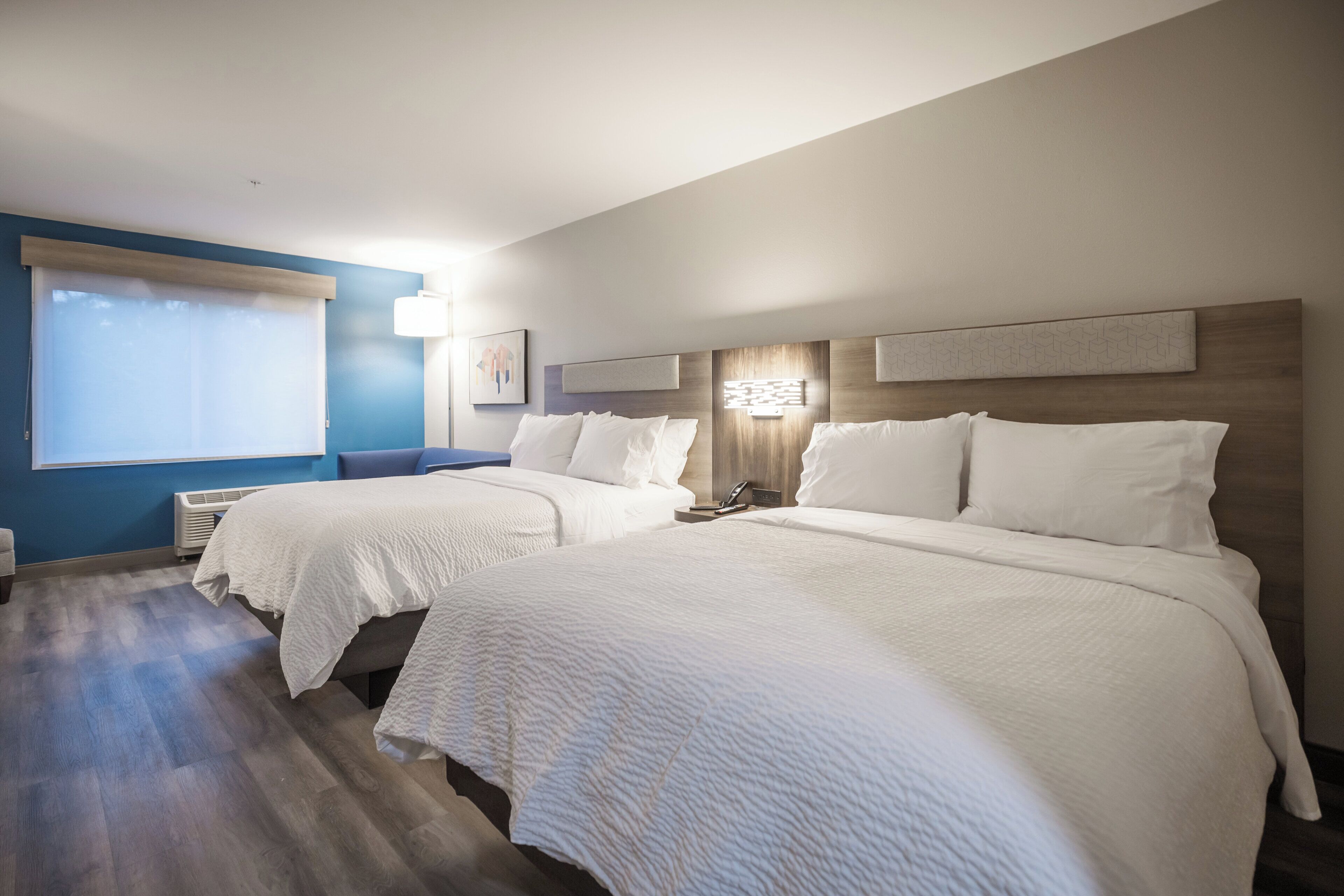 Ein Hotelzimmer mit zwei Betten, einem großen Fenster mit Jalousien, einer Wand-Klimaanlage und einem Nachttisch mit einer Lampe.
