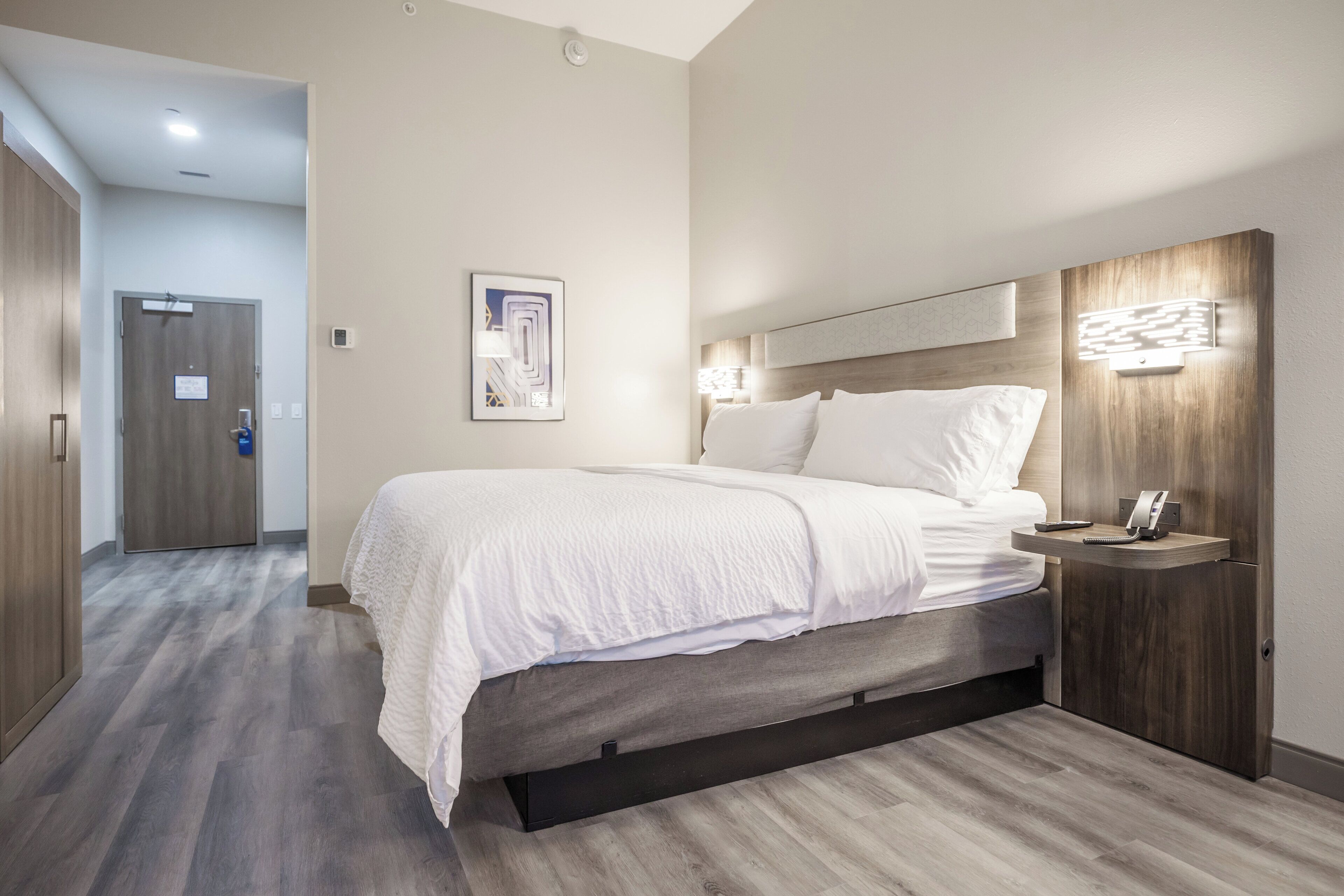 A modern hotel room with a large bed, a bedside table with a phone, and a wall-mounted light fixture.