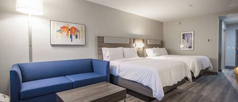 A hotel room with a blue sofa, a wooden coffee table, two beds with white bedding, and artwork on the walls.