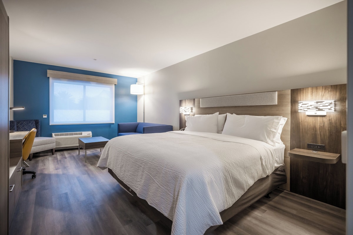 A modern hotel room with a large bed, a seating area with a sofa and armchair, a desk, and a window with blinds.