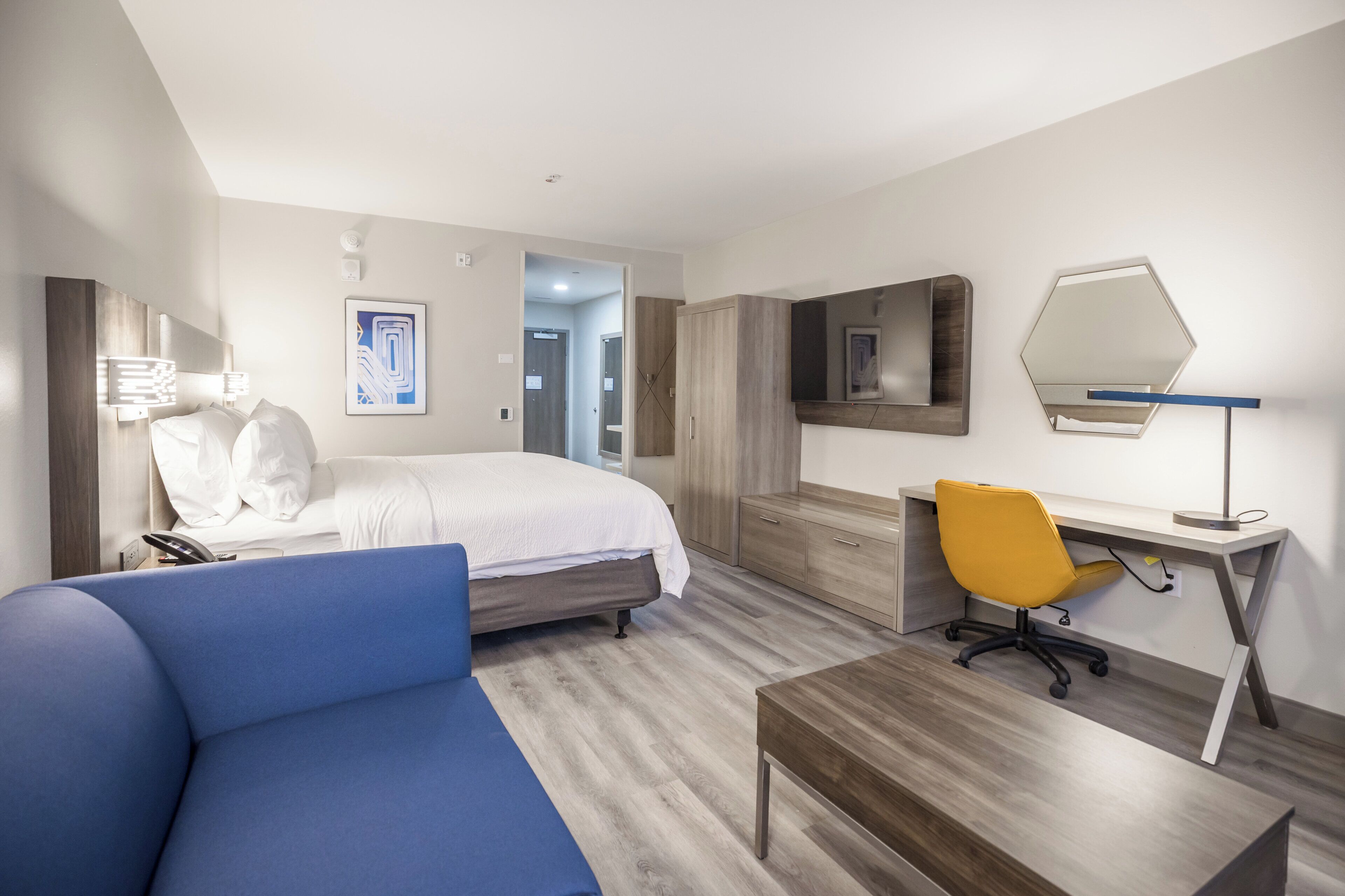 A hotel room with a bed, a desk, a chair, a mirror, and a blue sofa.