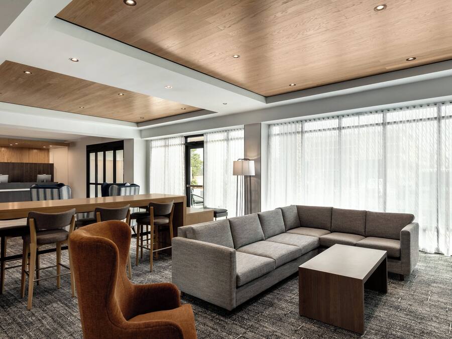 A modern hotel room with a sectional sofa, a wooden coffee table, a dining area with a table and chairs, and large windows with sheer curtains.