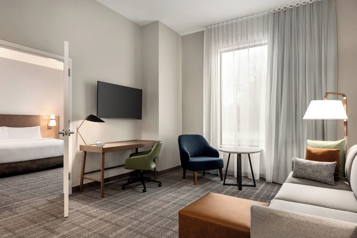 Une chambre d’hôtel comprenant un lit, un bureau, deux chaises, un canapé, une télévision et une fenêtre avec des rideaux.