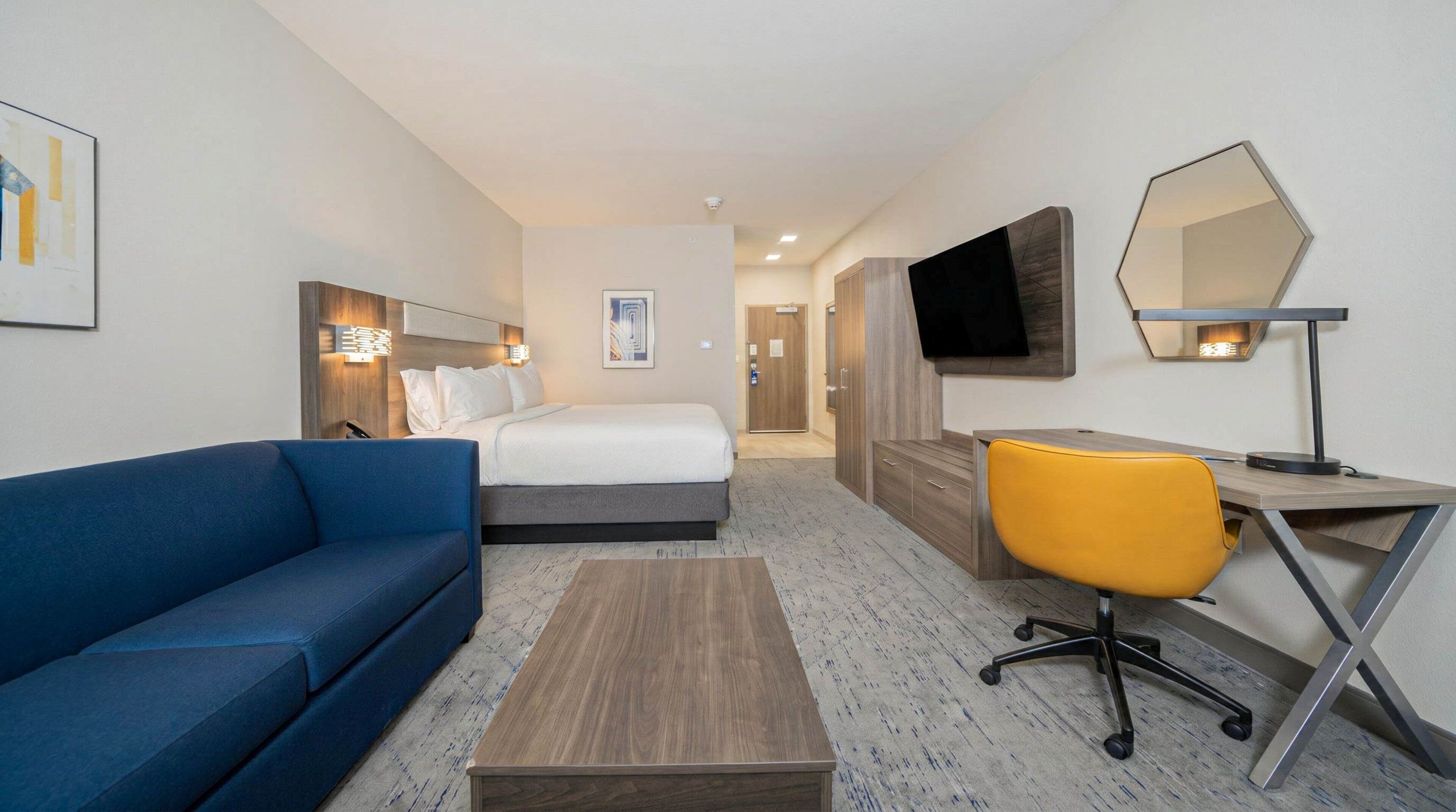 Hotellihuone, jossa on sänky, sininen sohva, työpöytä tuolilla, televisio ja peili.