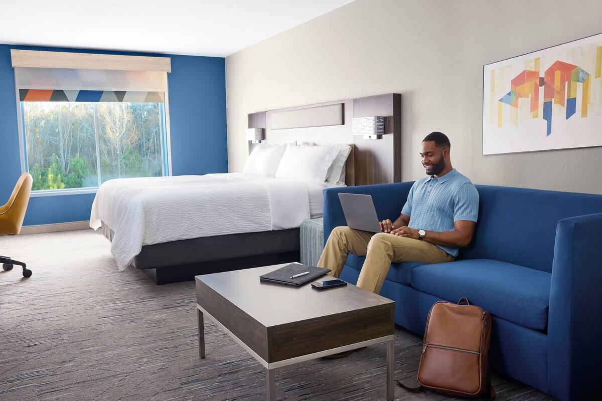 A hotel room with a bed, a blue sofa, a wooden coffee table, a laptop, a smartphone, and a leather bag.