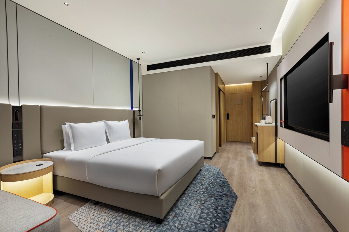 Una habitación de hotel moderna con una cama grande, una mesita de noche, un televisor y una pequeña kitchenette.
