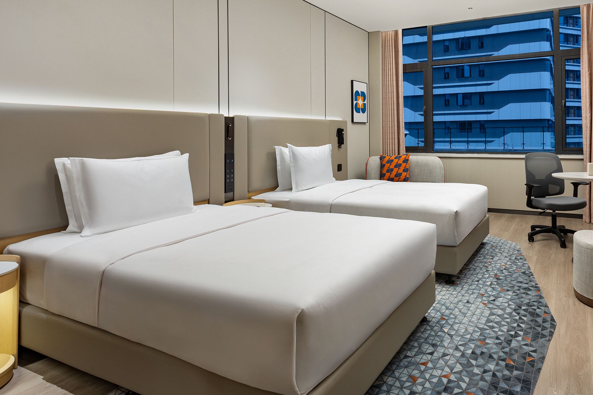 A hotel room with two beds, a desk, and a view of a high-rise building.