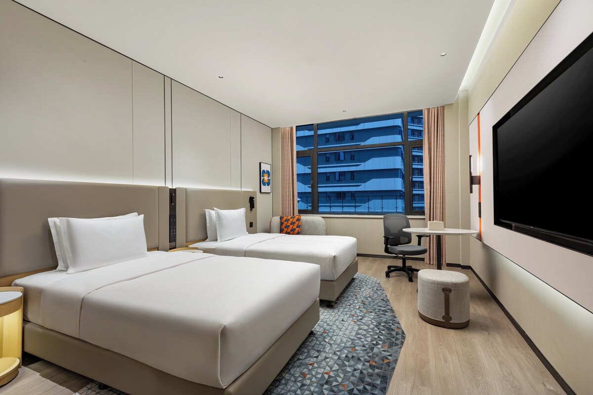 Habitación de hotel con dos camas, televisión, escritorio y ventana con vista a un edificio.