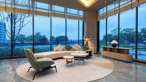 Un área de estar moderna con un sofá curvo, una mesa de centro redonda y una consola de madera con una lámpara. Amplias ventanas ofrecen vistas al exterior.