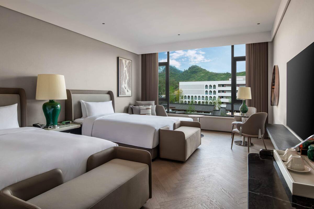 Ett hotellrum med två sängar, ett skrivbord, en stol och utsikt över en byggnad och berg.