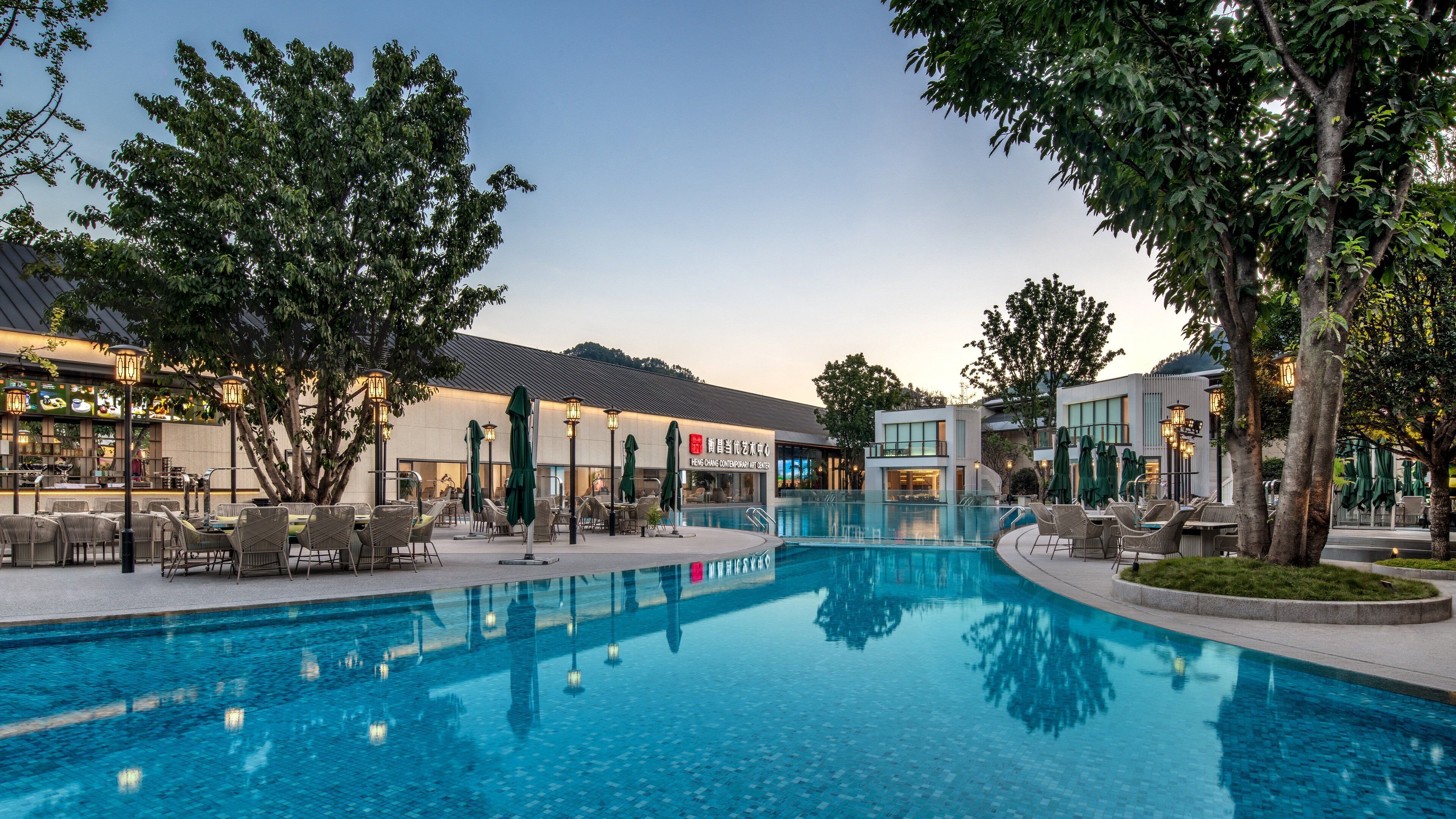 A swimming pool with lounge chairs and umbrellas, surrounded by trees and buildings with signage.