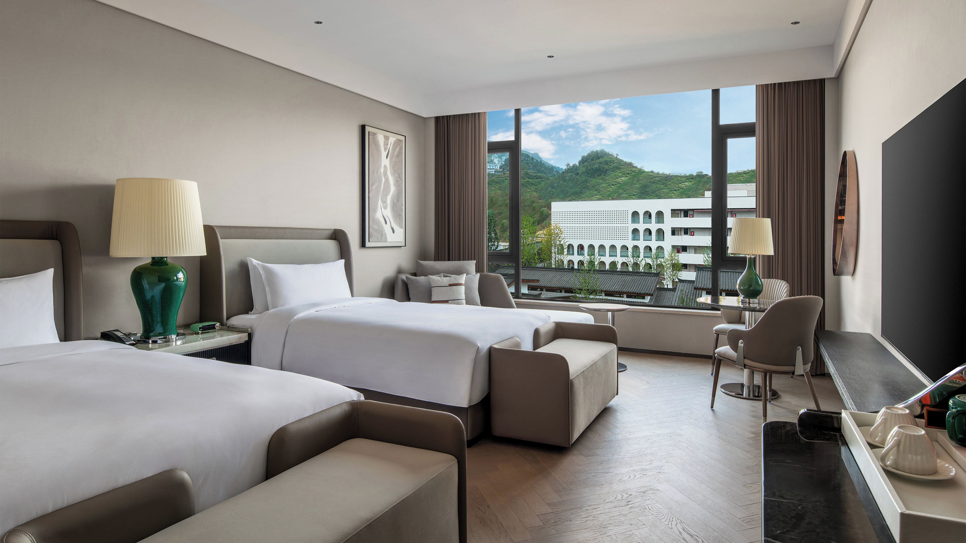 A hotel room with two beds, a sitting area, a desk, and a view of a building and greenery.