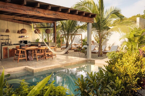 Un espace aménagé au bord de la piscine, comprenant une pergola en bois, une table à manger avec des chaises, un hamac et des fauteuils de relaxation.