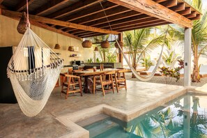 A poolside area with a wooden table and chairs, a hanging hammock, and a view of palm trees.