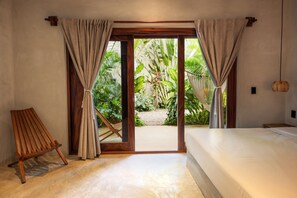A bedroom with a bed, wooden chair, and a view of a tropical garden through a glass door.
