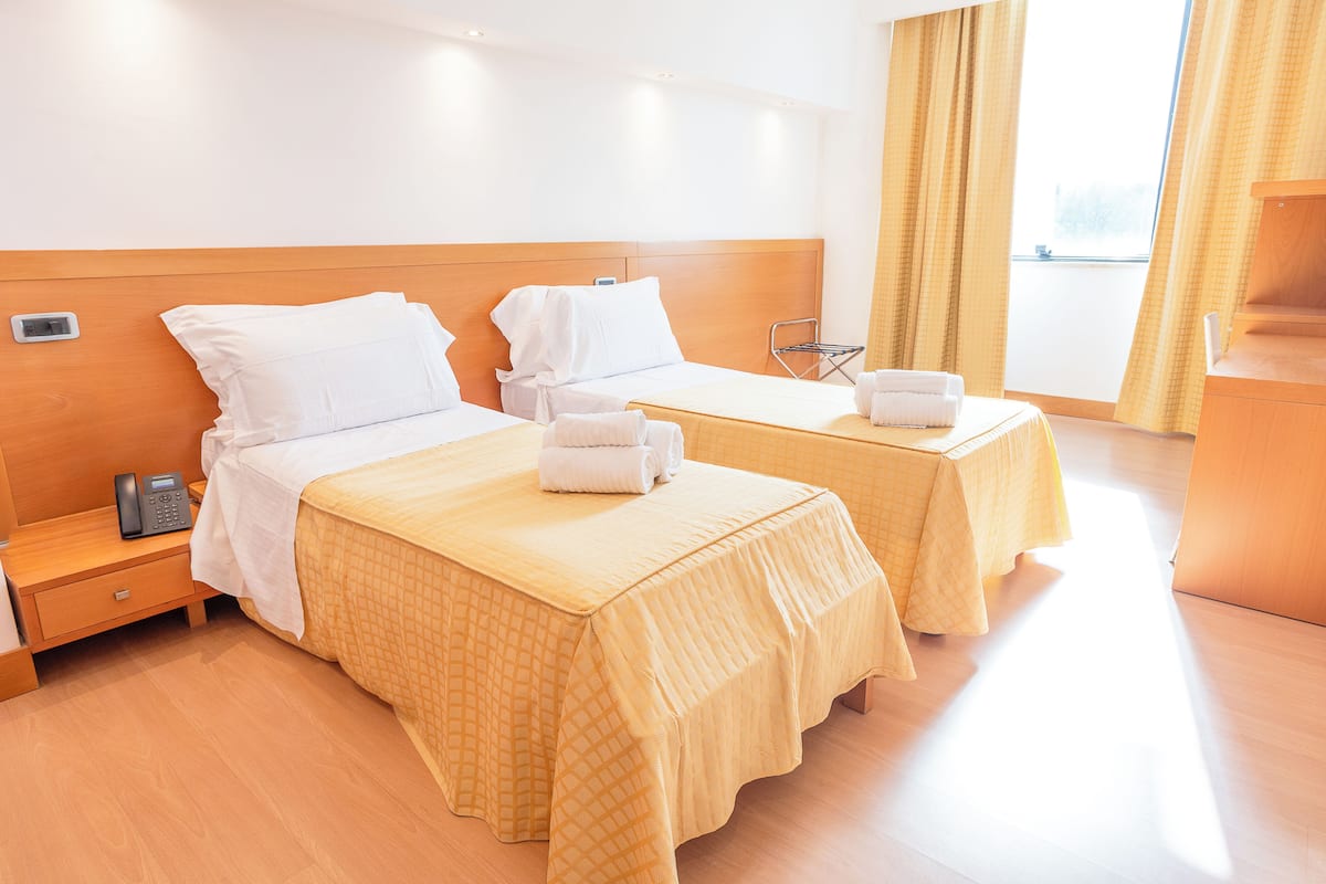A hotel room with two beds, a wooden headboard, a bedside table with a phone, and a window with curtains.