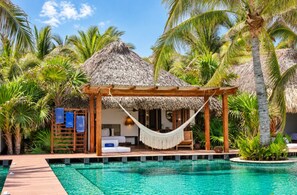 A poolside area with a thatched-roof structure, a hammock, and a bed with a view of the pool.