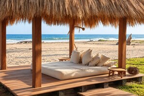 A beachside lounge with a thatched roof, wooden deck, and a cushioned seating area with pillows.