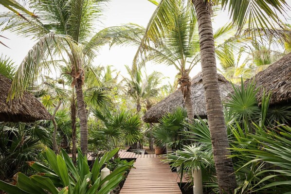 A wooden pathway surrounded by palm trees and thatched-roof huts in a tropical setting.